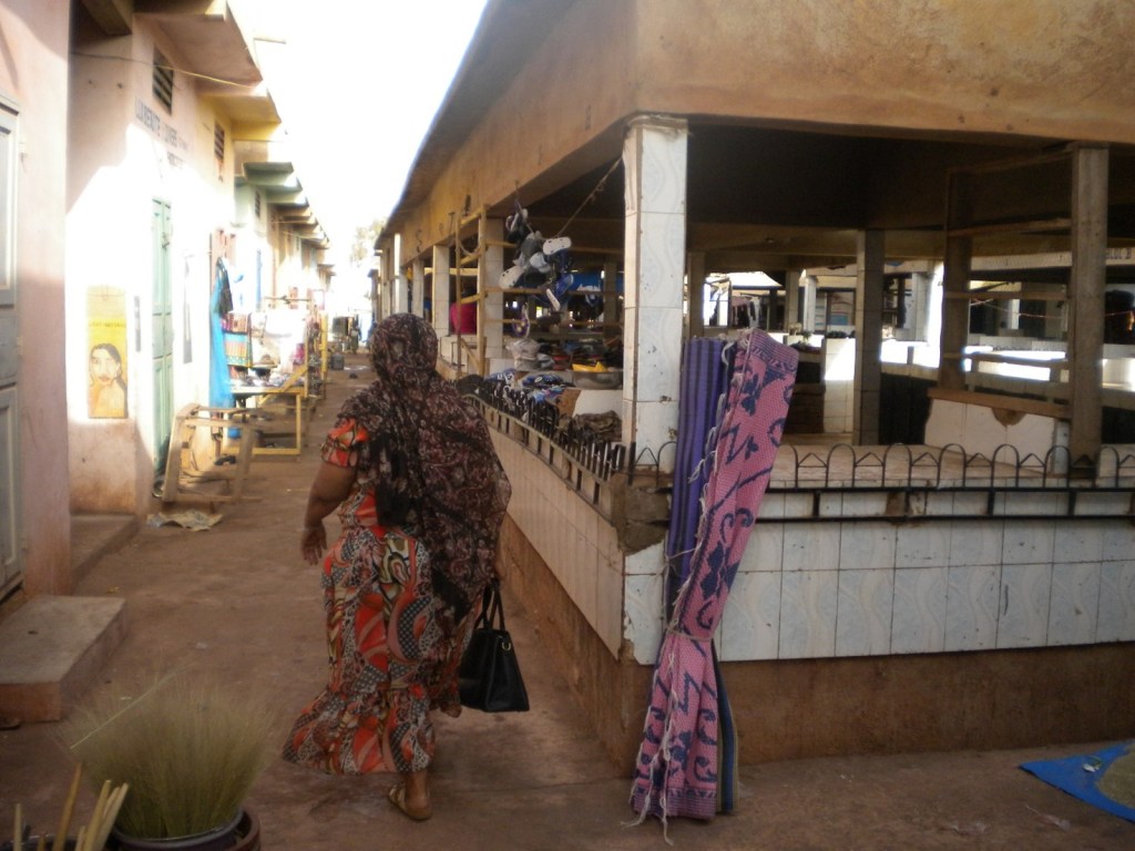 A woman dressed in highly patterned clothing, with her back to the camera, walks along a quiet lane in an urban market, bordered by small stalls in permanent buildings.