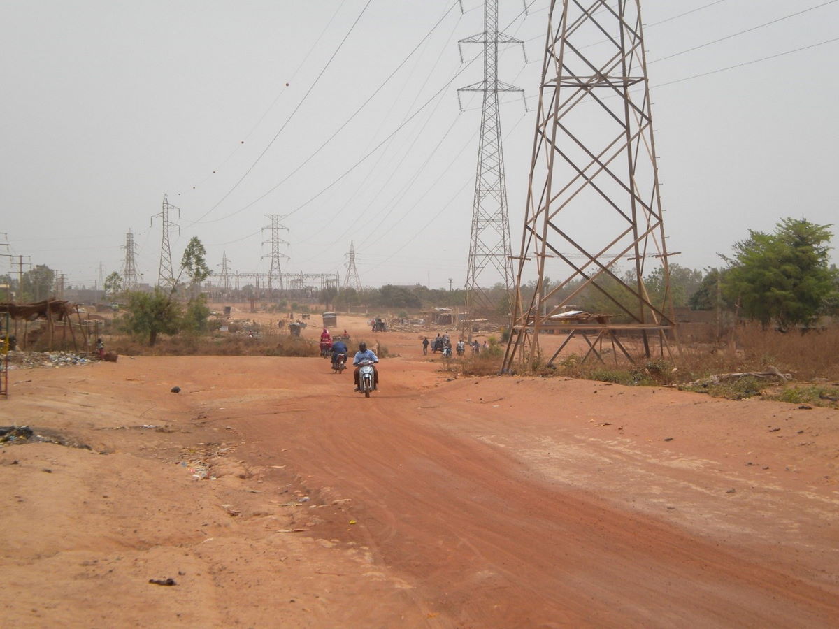 Motorbikes travel down a well-used, unmetalled, dusty road with sandy verges, bordered by trees and a chain of many electricity pylons